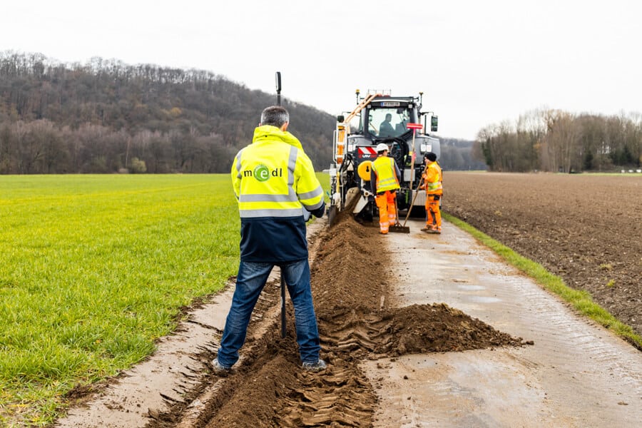 medl-Mitarbeiter dokumentiert in der Saarner Ruhraue die frisch verlegte Glasfaserleitung am Graben, den die im Hintergrund erkennbare Baumaschine, eine sog. Layjet-Verlegeeinheit, eben geschlossen hat.