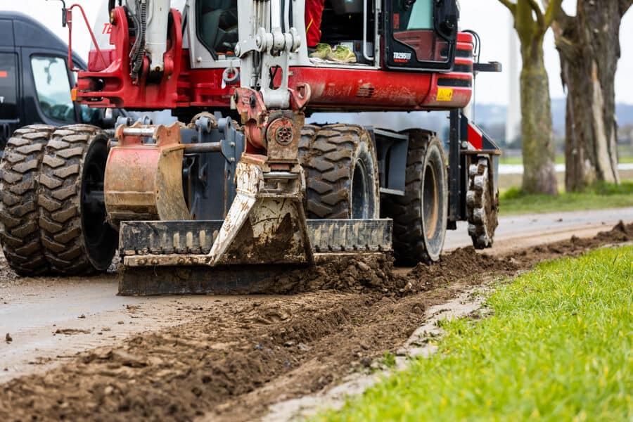 Nahaufnahme eines Baggers, die Trasse nach dem Glasfaserausbau mit Layjet-Technologie im Randbereich einer Straße in der Saarner Ruhraue verdichtet und säubert.
