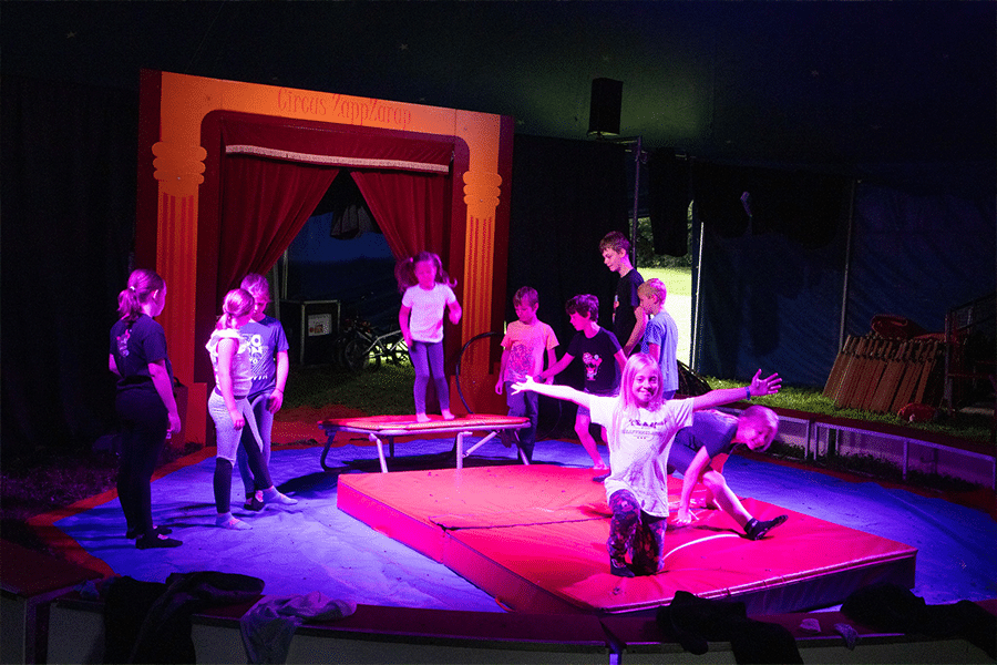 Eine Gruppe von Kindern übt in der Manege des medl-Mitmach-Zirkus' Sprünge von einem kleinen Trampolin auf eine große Matte.