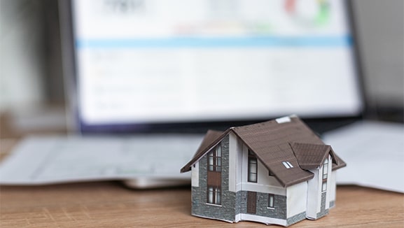 Fernwaerme-Finanzierungen_577x326 Foto von einem Miniaturhaus auf einem Tisch. Im Hintergrund sind unscharf Zettel und ein Bildschirm mit Diagrammen und Tabellen zu erkennen. Symbolbild für Förderungen von energetischen Gebäudesanierungen.