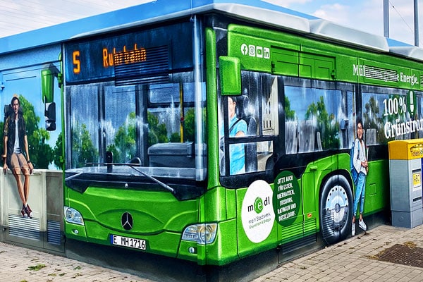 Ein bemaltes Gebäude, eine sog. Trafostation, mit einer künstlerischen Darstellung eines Mülheimer Busses gestaltet im medl-Design an einer Haltestelle. Im Hintergrund sitzt eine Person auf einer Mauer im Vordergrund steht eine wartende Frau neben einem Ticketautomaten