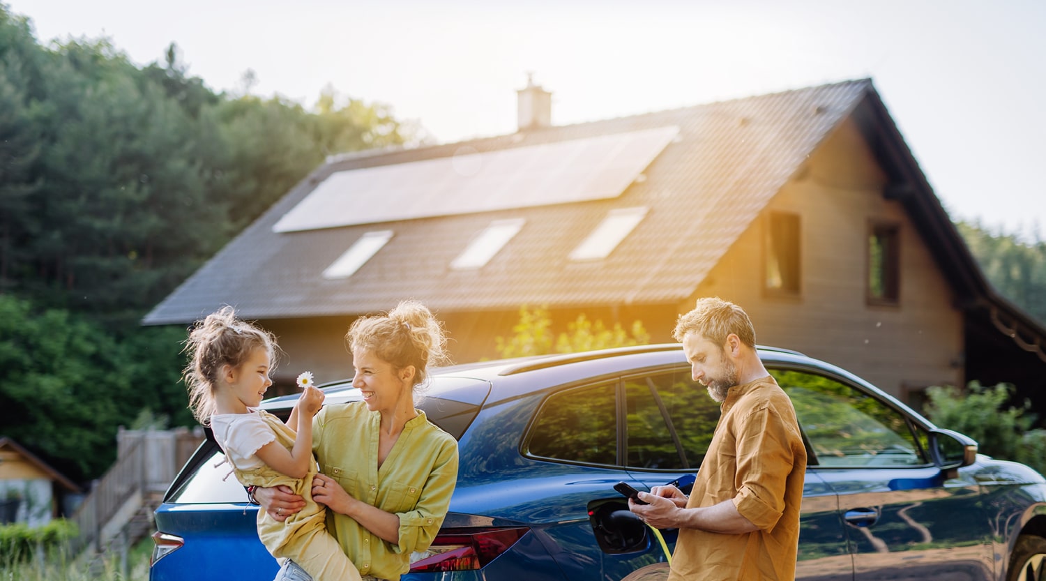 Ein Mann lädt ein Auto vor einem Haus mit Solarmodulen auf dem Dach, daneben steht eine Frau mit Kind auf dem Arm.