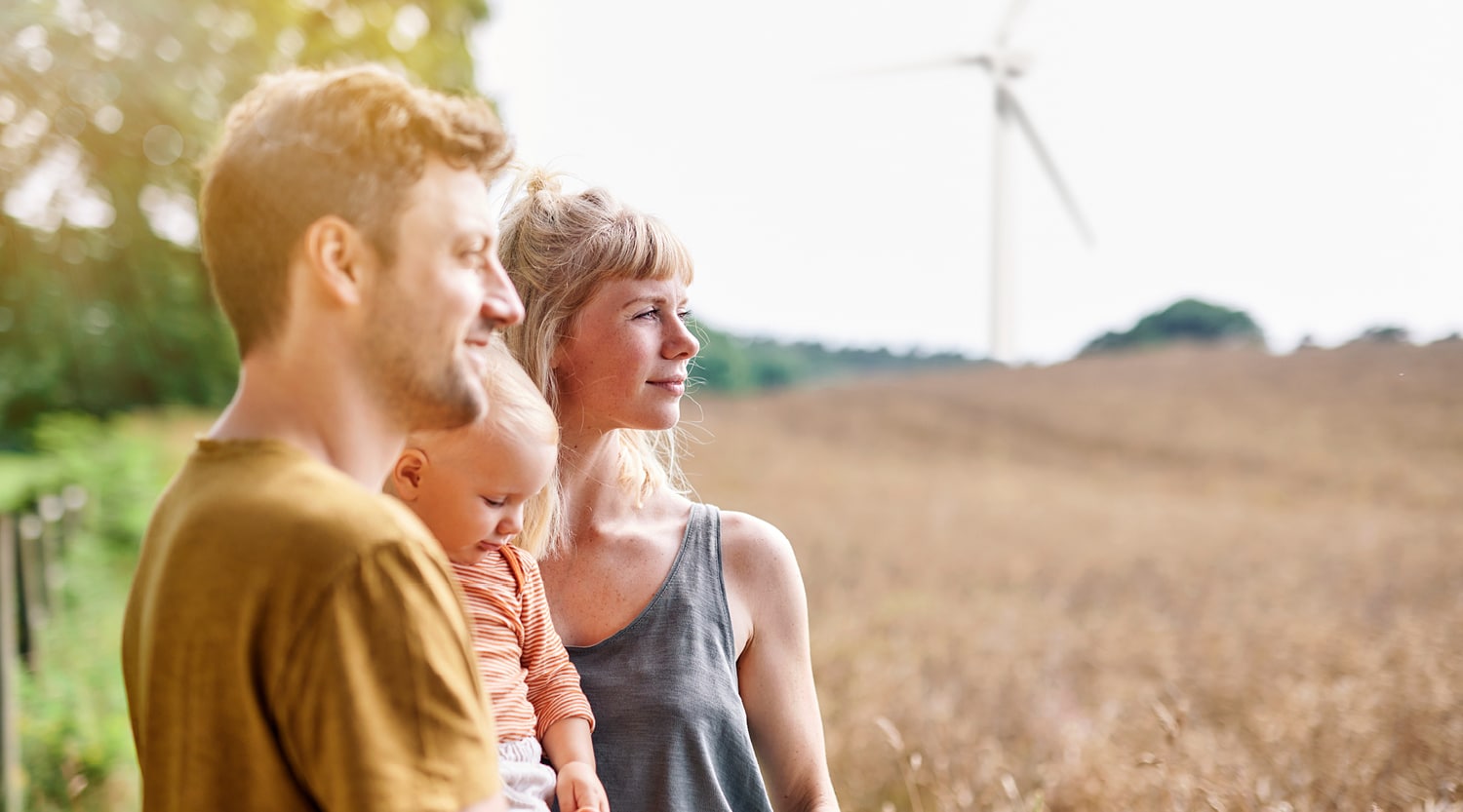 Drei Personen stehen auf einem Feld, ein Kind wird getragen, im Hintergrund steht eine Windenergieanlage.