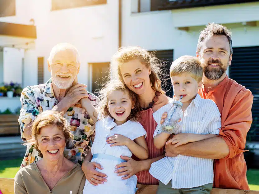 Zuhause laden als Wohnungseigentümer: Lachende Familie vor einem Mehrfamilienhaus – Symbol für zufriedenen Wohnungseigentümer unterschiedlichen Alters.