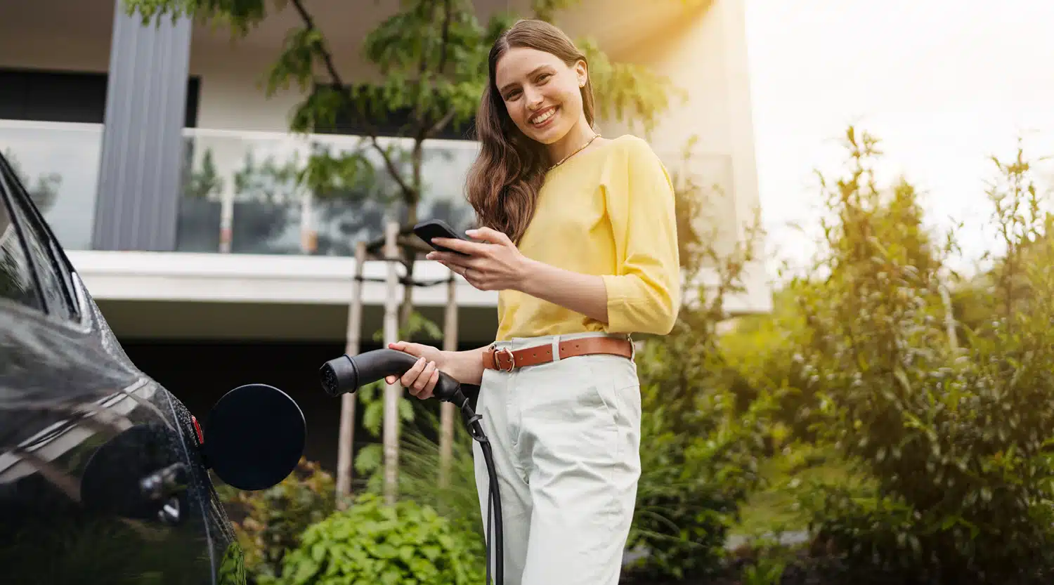 Lächelnde Frau steht vor einem Mehrfamilienhaus mit einem Stecker in der Hand an ihrem eAuto – Symbol für das zu Hause laden als Wohnungseigentümer