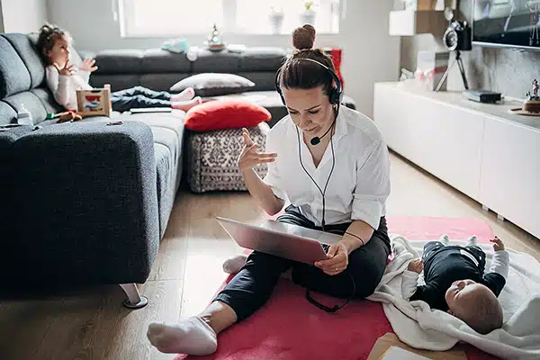 Eine Frau mit Headset arbeitet gestikulierend am Laptop auf dem Wohnzimmerboden, neben ihr ein Baby, während ein kleines Mädchen im Hintergrund auf der Couch fernsieht – perfektes Internet mit GigaGlasfaser für die ganze Familie.