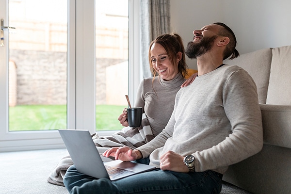 Ein lächelndes Paar sitzt auf dem Boden vor einem Laptop in einem modernen Wohnzimmer – und genießen freuen sich über die GigaGlasfaser-Vorzugspreise für SWB-Mieter.