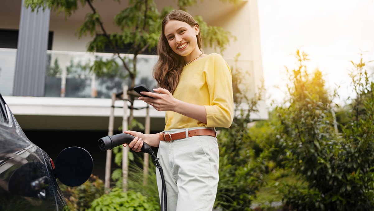 Lächelnde Frau lädt E-Auto vor Wohnhaus und hält Smartphone, Symbolbild für einfache medl Ladelösungen.