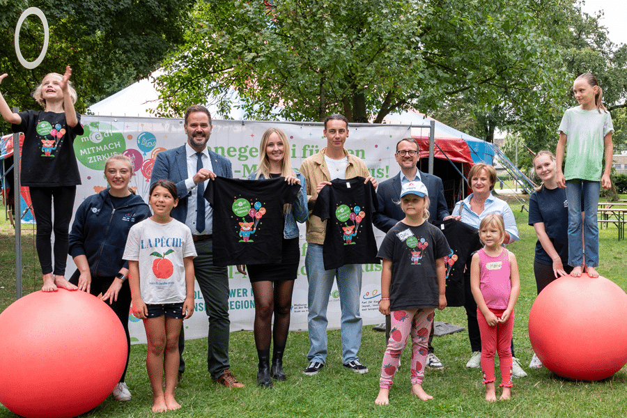 Gruppenfoto beim medl-MITMACH Zirkus: Kinder und Erwachsene posieren im Stadtpark, einige balancieren auf roten Gymnastikbällen, andere halten schwarze T-Shirts mit Zirkusmotiv hoch.