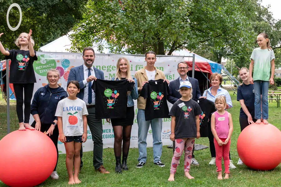 Gruppenfoto beim medl-MITMACH Zirkus: Kinder und Erwachsene posieren im Stadtpark, einige balancieren auf roten Gymnastikbällen, andere halten schwarze T-Shirts mit Zirkusmotiv hoch.
