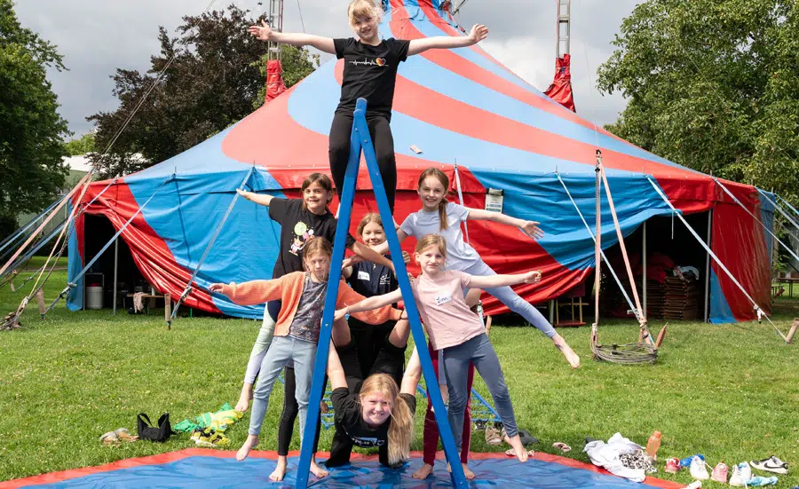 Kinder trainieren beim medl-mitmach-zirkus vor einem rot-blauen Zirkuszelt und bilden gemeinsam eine akrobatische Pyramide auf der Wiese.