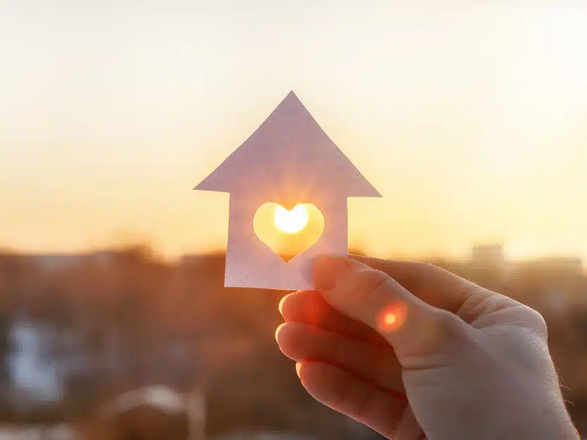 Hand hält Papier-Haus mit Herz-Ausschnitt in die Sonne – Symbol für Wertsteigerung der Immobilie durch Photovoltaik.