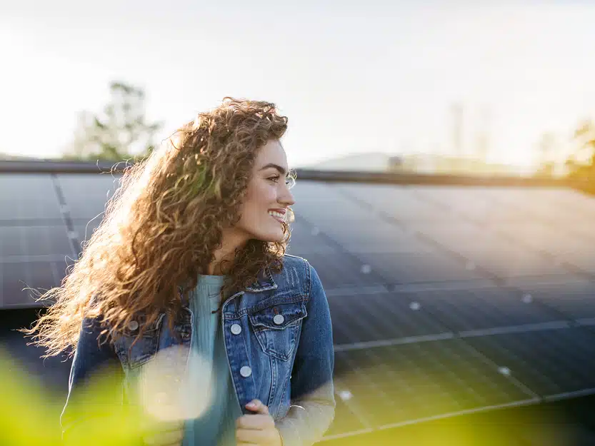 Junge Frau steht lächelnd vor Solarmodulen auf dem Dach und blickt in die Sonne – Symbolbild für Klimaschutz durch Photovoltaik.