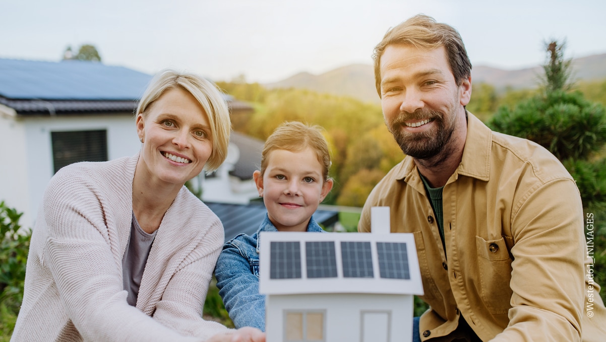 Glückliche Familie hält Modellhaus mit Solaranlage in die Kamera, im Hintergrund echtes Haus mit PV-Modulen.