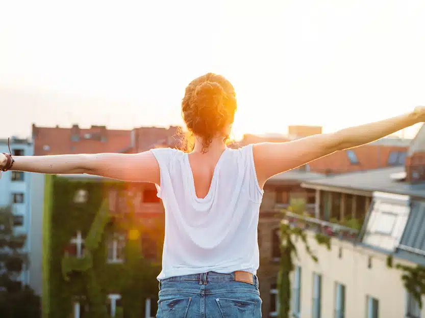 Frau mit ausgebreiteten Armen auf Dachterrasse blickt in die Sonne, symbolisiert Unabhängigkeit durch Photovoltaik.
