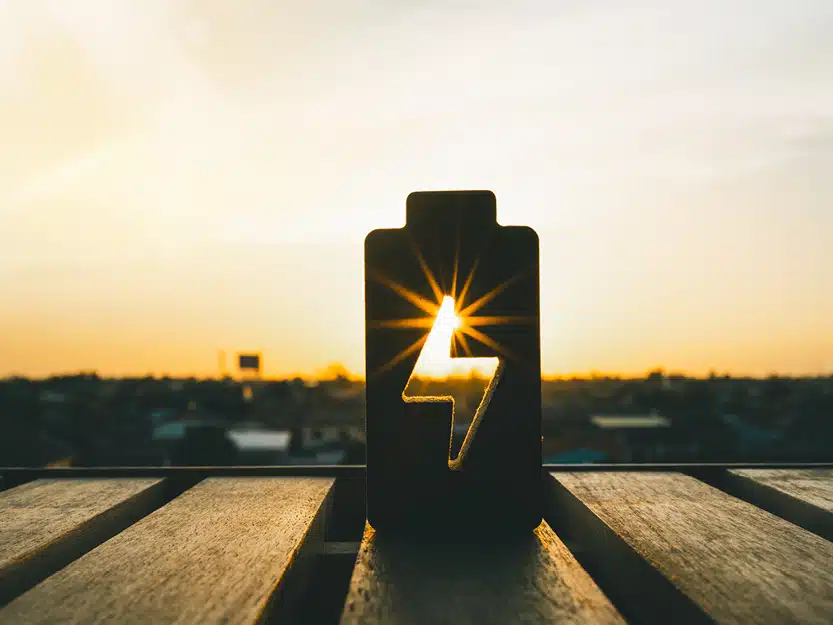 Batteriemodell in der Abenddämmerung auf einem Holztisch mit ausgeschnittenem Blitzsymbol in der Mitte, durch das die untergehende Sonne scheint – Symbol für nachhaltiges Laden als Wohnungseigentümer