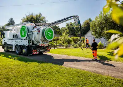 Mitarbeiter der sem GmbH bei der Reinigung einer Kleinkläranlage in einer Mülheimer Schrebergartenanlage. Zu sehen ist das spezialisierte Reinigungsfahrzeug sowie das geschulte Personal bei der fachgerechten Wartung der Anlage in entsprechender Arbeitskleidung. Im Hintergrund sind Gartenparzellen erkennbar.