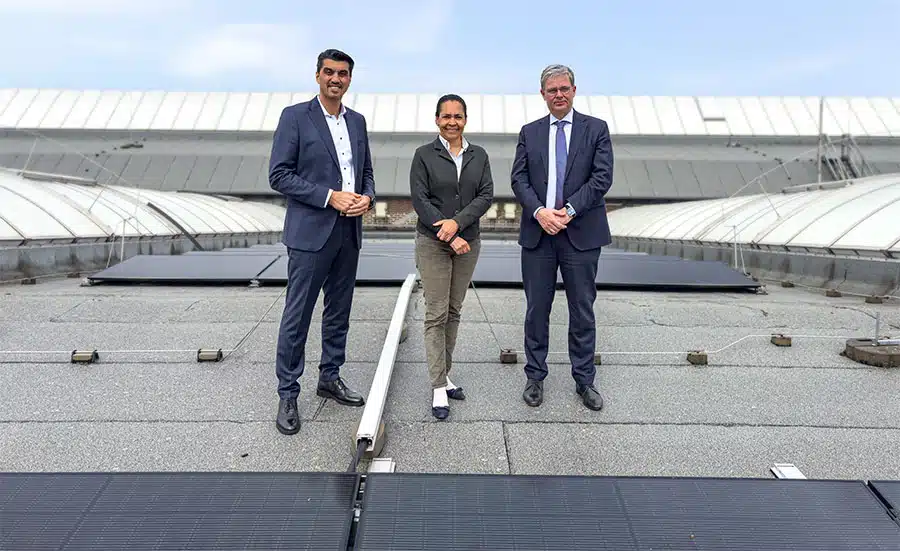 Von medl gebaute Photovoltaik-Anlage auf dem Dach der Ruhrbahn in Mülheim Drei Personen in Business-Kleidung stehen lächelnd auf einem Flachdach. Im Vordergrund liegen flache, schwarze Photovoltaik-Module. Im Hintergrund sind große, gebogene Hallendächer des Ruhrbahn-Betriebshofs in Mülheim zu sehen. Der Himmel ist leicht bewölkt.