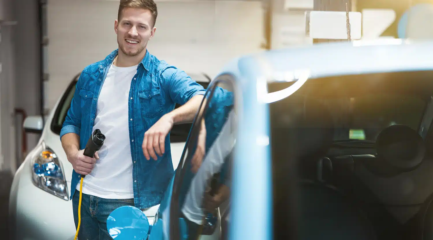 Lächelnder Mann mit Ladestecker in der Hand lehnt lässig an einem Auto in einer Garage – Symbol für das Laden in der Firma