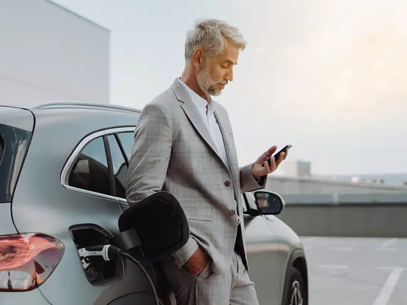 Frau lehnt lässig an seinem Firmenwage und schaut aufs Handy, während seub Elektroauto an der heimischen Wallbox lädt – Symbol für transparente Kosten beim Laden zu Hause.