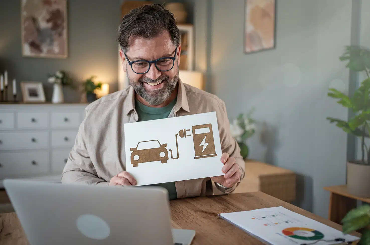 Lächelnder Mann sitzt in seinem Zuhause mit einer Grafik von einem ladenden eAuto in der Hand und schaut auf seinen Computerbildschirm – Symbol für die Unterstützung bei der Anschaffung der privaten Ladeinfrastruktur